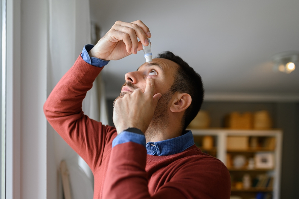 A man in a red sweater applies eye drops while looking up - Optometrist Chapel Hill A man in a red sweater applies eye drops while looking up - Optometrist Chapel Hill