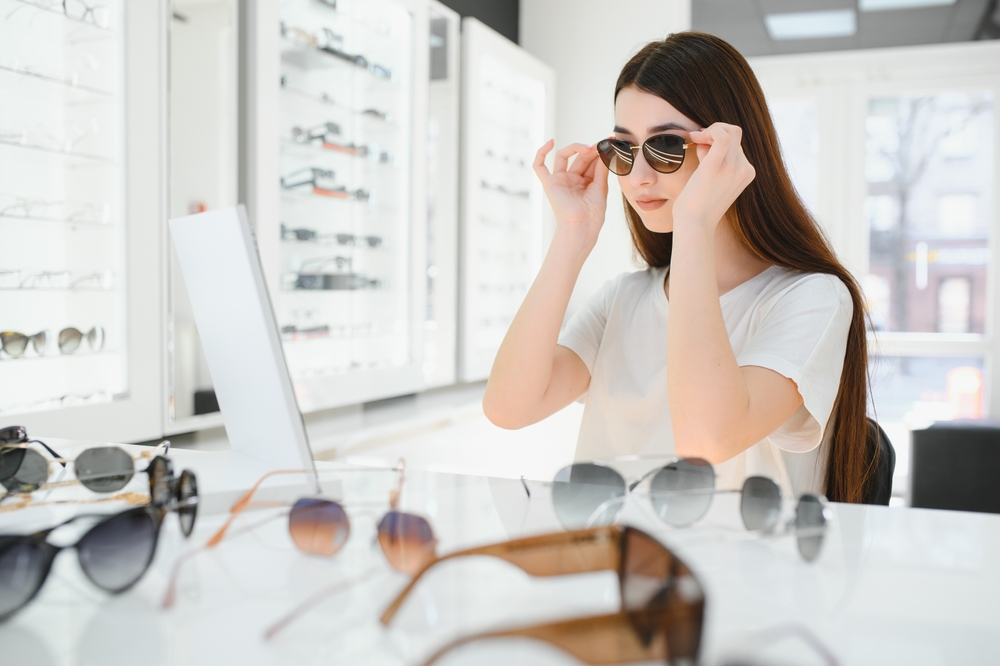 A woman tries on sunglasses at an eyewear store, surrounded by various pairs on display - Optometrist Chapel Hill A woman tries on sunglasses at an eyewear store, surrounded by various pairs on display - Optometrist Chapel Hill