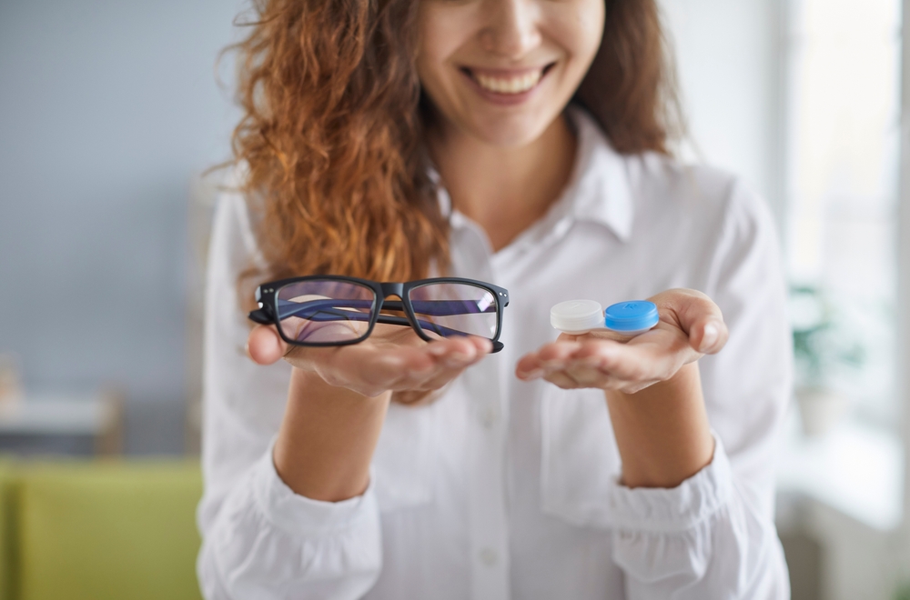 A smiling woman holds a pair of glasses in one hand and a contact lens case in the other - Optometrist Chapel Hill A smiling woman holds a pair of glasses in one hand and a contact lens case in the other - Optometrist Chapel Hill