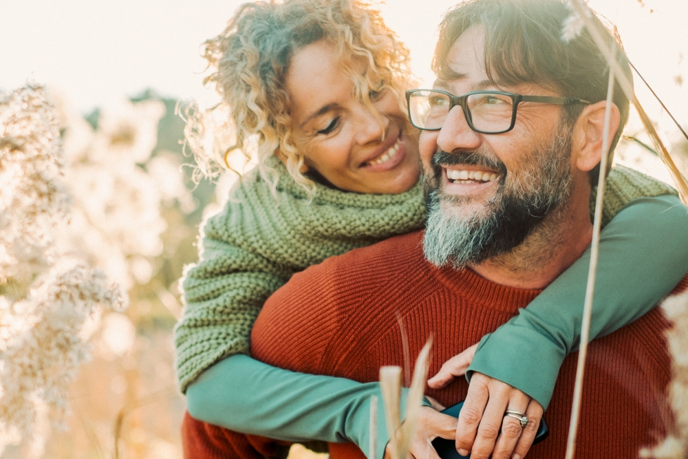 A smiling woman embraces a man from behind, both wearing cozy sweaters and enjoying a sunny outdoor moment - Optometrist Chapel Hill A smiling woman embraces a man from behind, both wearing cozy sweaters and enjoying a sunny outdoor moment - Optometrist Chapel Hill