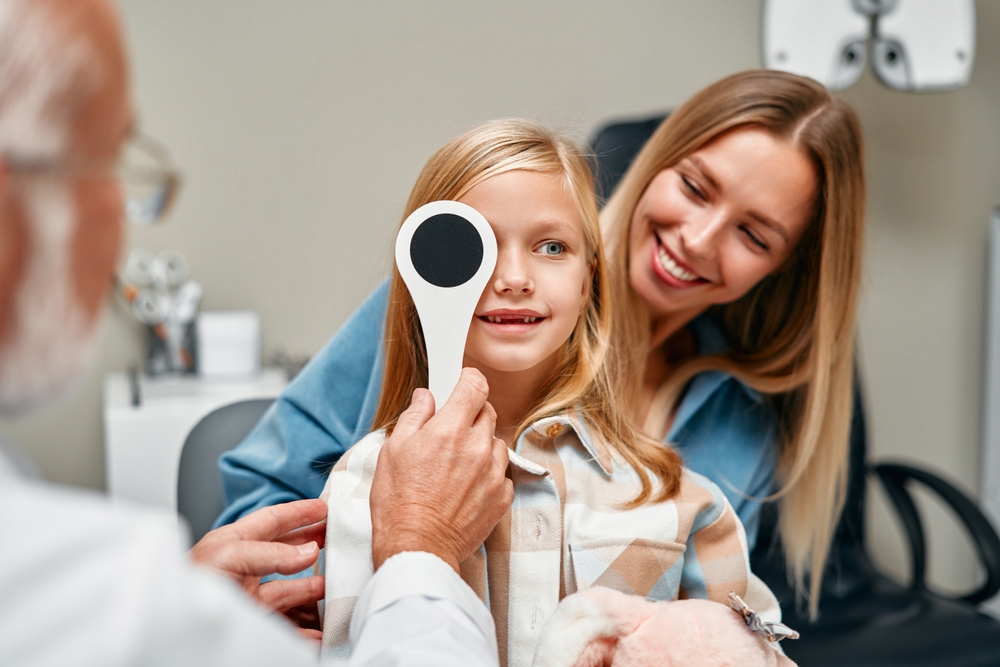 An optometrist performs an eye exam on a young girl while her mother smiles beside her - Optometrist Chapel Hill An optometrist performs an eye exam on a young girl while her mother smiles beside her - Optometrist Chapel Hill