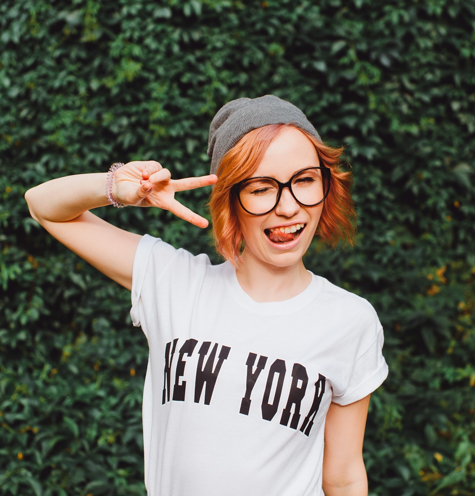 A young woman wearing glasses, a beanie, and a "New York" shirt poses playfully while making a peace sign and sticking her tongue out - Optometrist Chapel Hill A young woman wearing glasses, a beanie, and a "New York" shirt poses playfully while making a peace sign and sticking her tongue out - Optometrist Chapel Hill