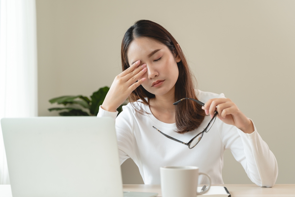 A woman sitting at a desk rubs her eyes while holding her glasses in one hand, appearing fatigued from working on a laptop - Optometrist Chapel Hill A woman sitting at a desk rubs her eyes while holding her glasses in one hand, appearing fatigued from working on a laptop - Optometrist Chapel Hill